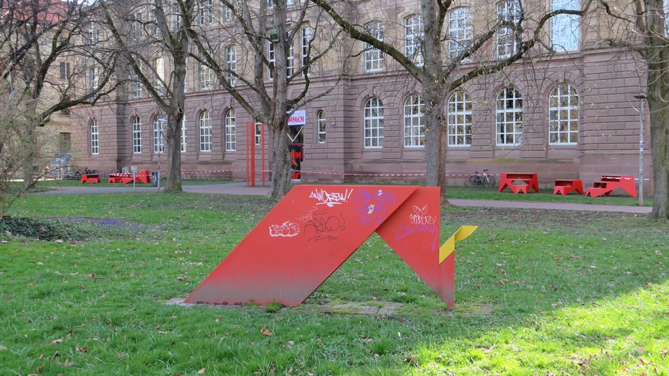 Im Vordergrund Kunstwerk "Der Schritt ins Freie" und im Hintergrund rote Sitzmöbel der Produktlinie STOOP vor Hauptgebäude der Hochschule