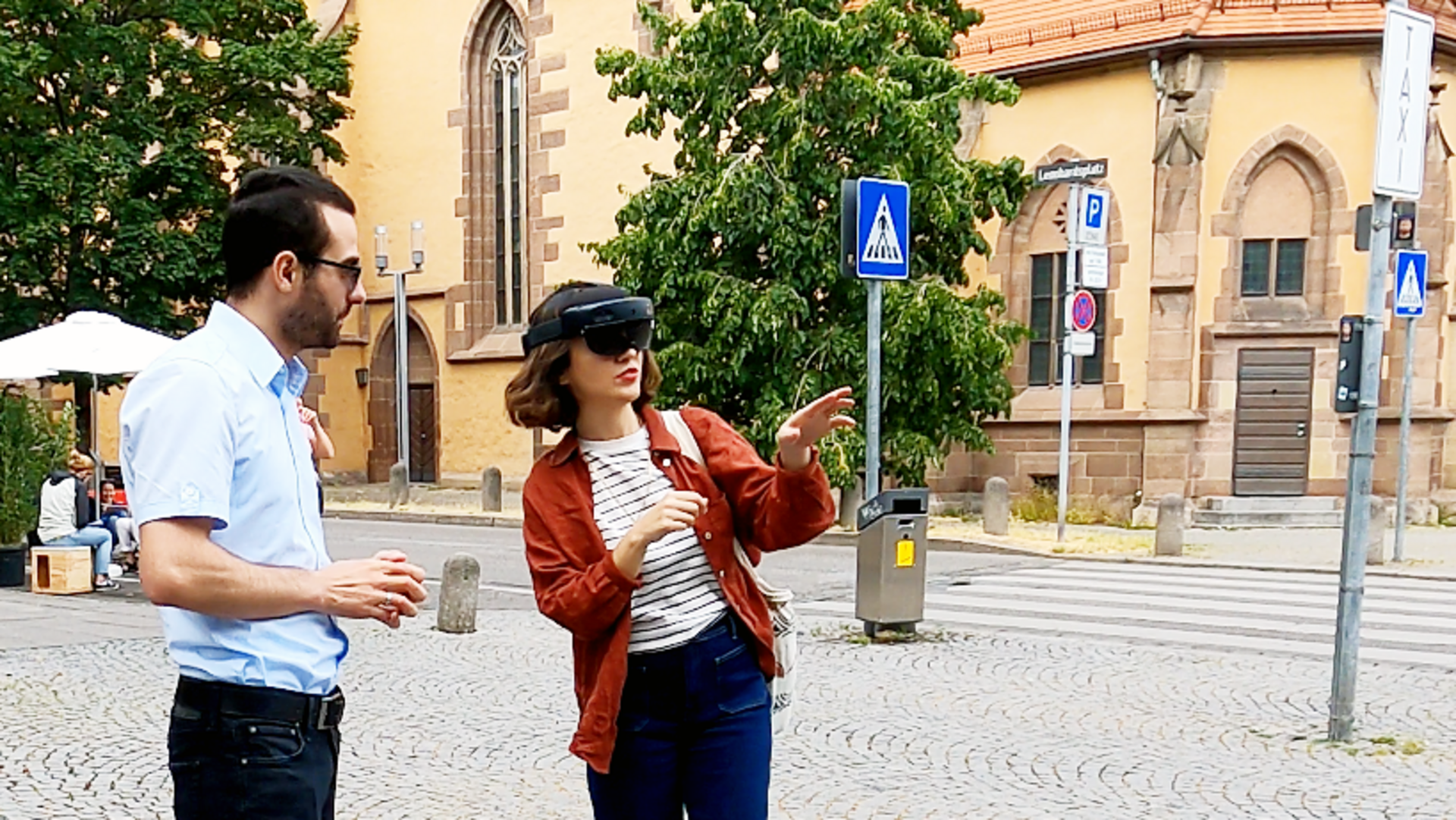 Eine Frau steht mit einer VR-Brille in der Leonhardsvorstadt und lässt sich von einem Mann die Anwendung erklären, im Hintergrund die Leonhardskirche