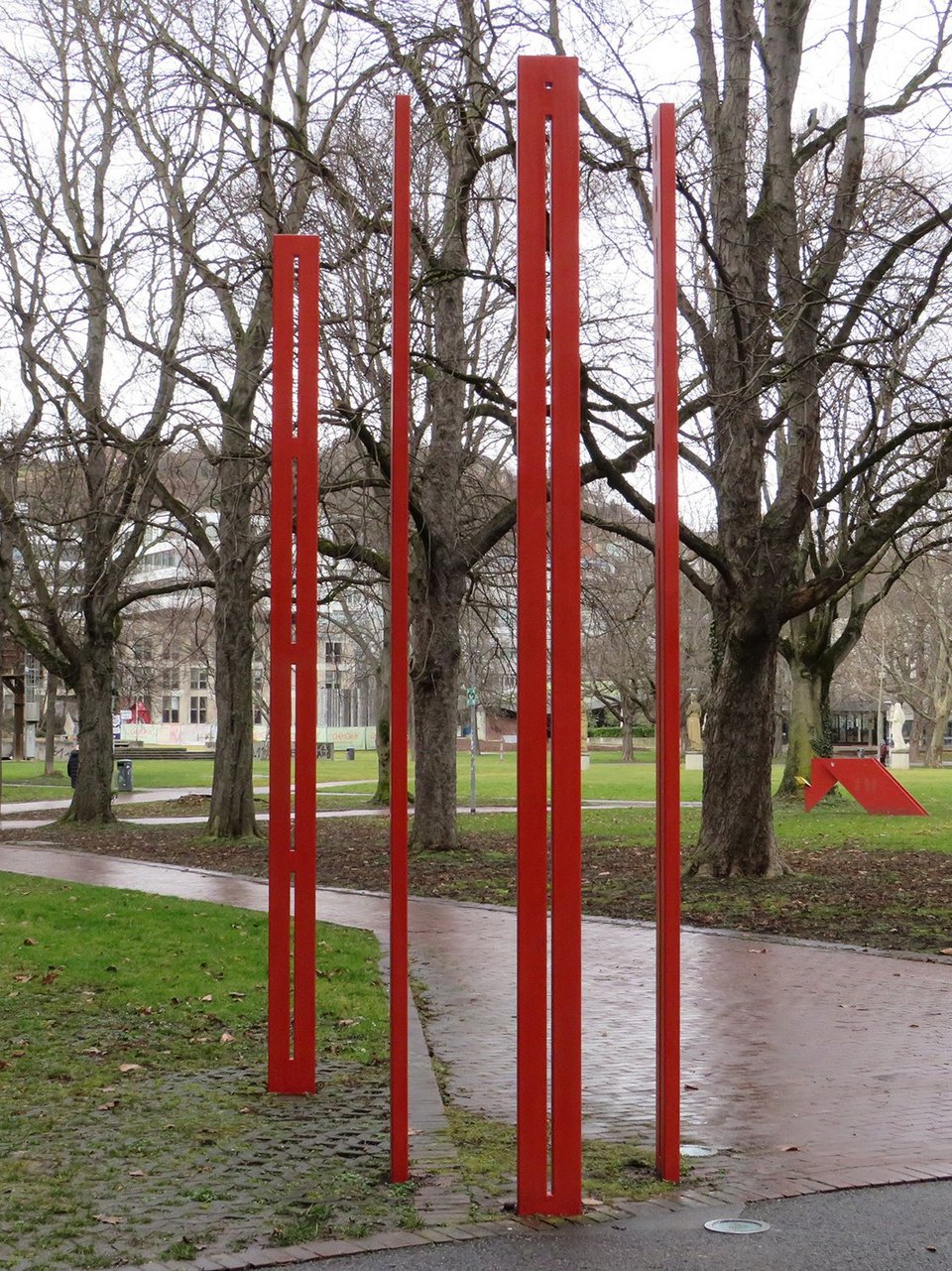 Vier rote hohe schmale Stahlstelen vor einer Parkanlage mit großen Bäumen.