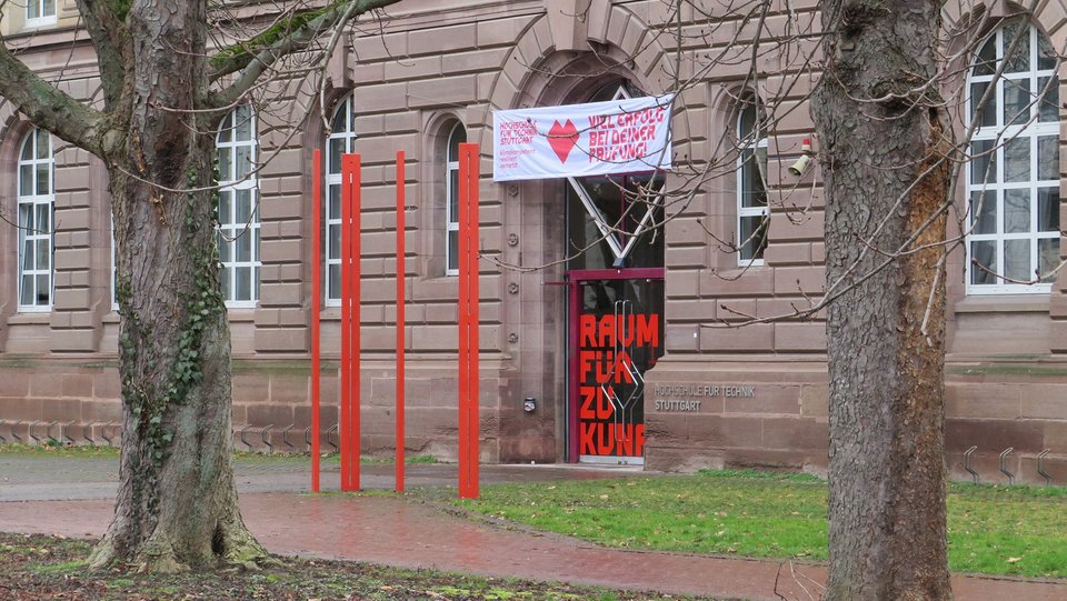 Vier rote Stelen vor dem Eingang des Hochschulgebäudes, zwischen zwei Bäumen hindurch fotografiert.