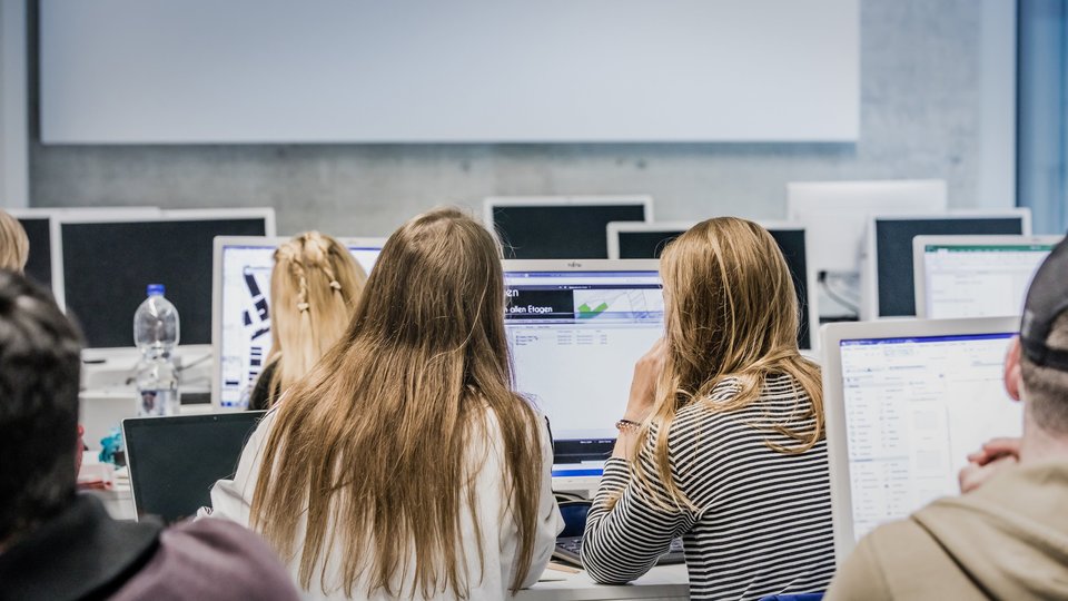 Auf dem Bild sind 2 Studierende während eienr Vorlesung zu sehen, die am Computer Aufgaben bearbeiten.